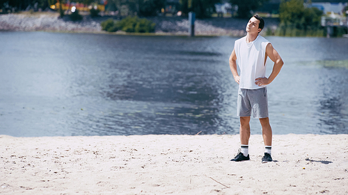 sportsman in tank top and shorts warming up neck and standing with hands on hips near river