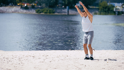 young sportsman in tank top and shorts warming up with outstretched hands near river