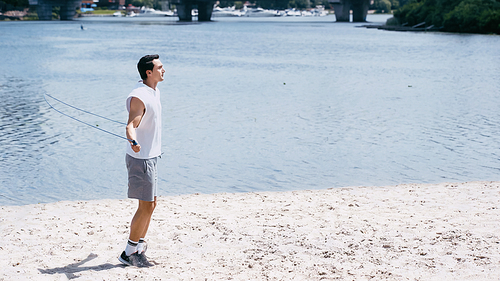 side view of young man jumping with skipping rope on sand near river