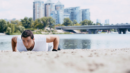 young sportsman doing push ups on sand near river in city