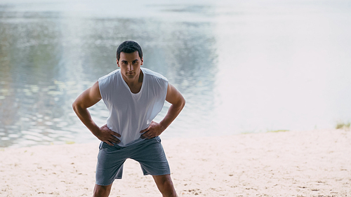 young sportsman in tank top working out with hands on hips near river