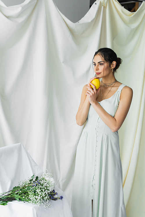 dreamy woman in dress holding lemon near bouquet of flowers on white