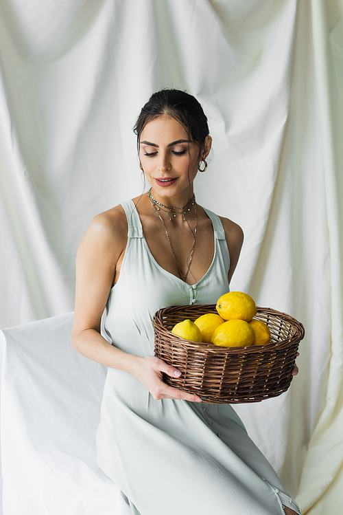 happy woman in dress holding wicker basket with lemons on white