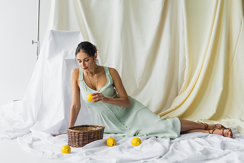 full length of pretty woman in dress holding lemon and looking at wicker basket on white