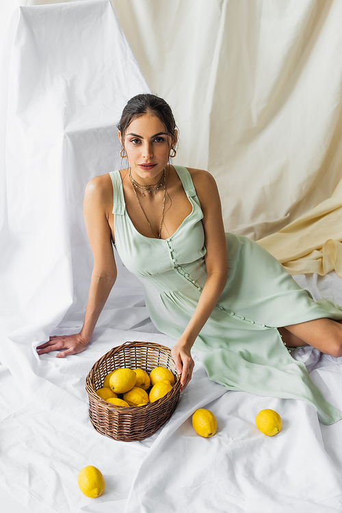 high angle view of pretty woman in dress sitting near wicker basket with lemons on white