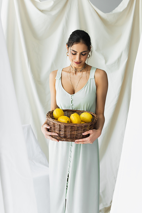 smiling woman in dress looking ripe lemons in wicker basket on white