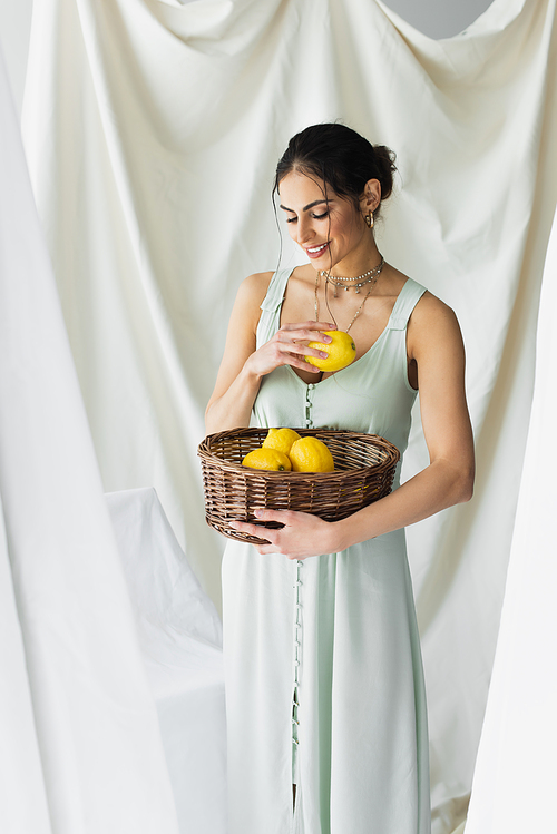 pleased woman in dress holding ripe lemons in wicker basket on white