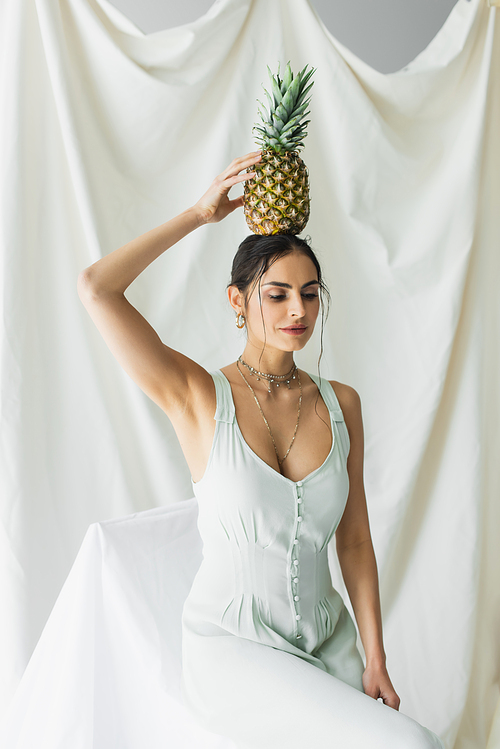 brunette woman in dress holding pineapple above head on white