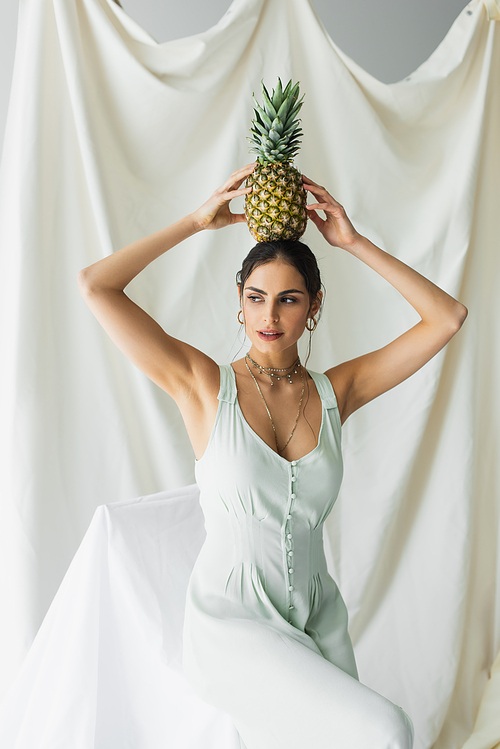 brunette woman in dress posing with pineapple above head on white