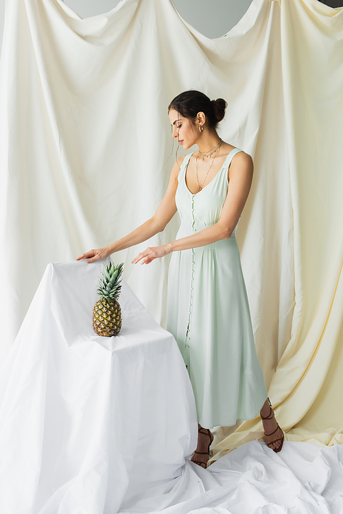 full length of brunette woman in dress posing near ripe pineapple on white