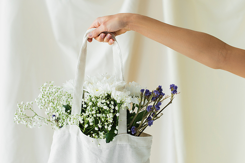 partial view of woman holding fabric shopper bag with flowers on white