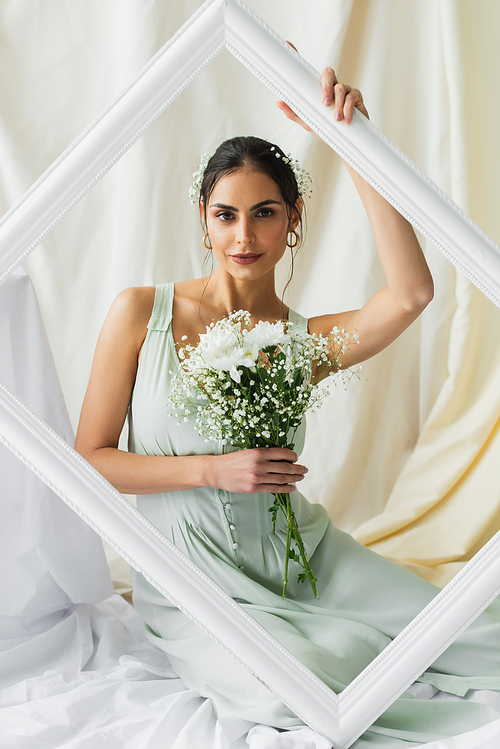 brunette woman holding bouquet of blooming flowers and posing near frame on white