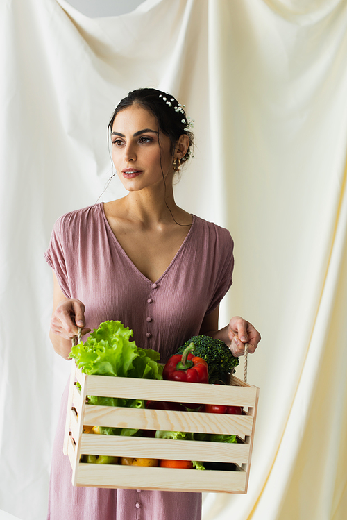 brunette woman holding wooden box with vegetables on white