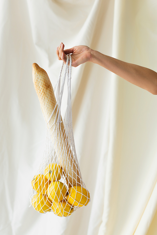 cropped view of woman holding reusable string bag with lemons and baguette on white