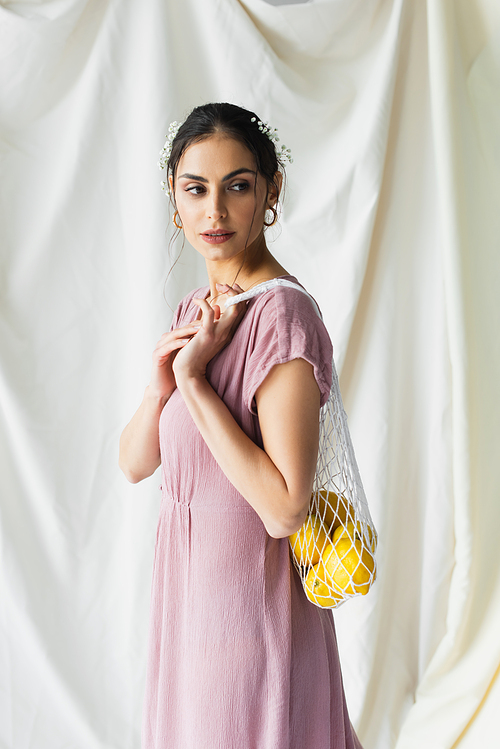 brunette woman holding reusable string bag with lemons on white