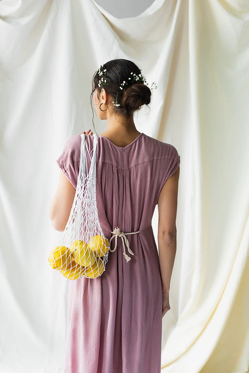 back view of brunette woman holding reusable string bag with lemons on white