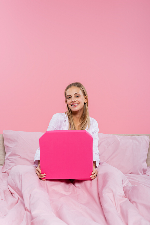 Happy blonde woman holding pizza box on bed isolated on pink