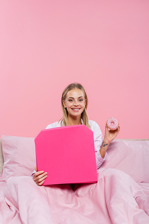 Smiling blonde woman in pajamas holding donut and pizza box on bed isolated on pink