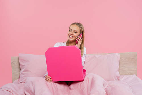 Smiling woman in pajamas talking on smartphone and holding pizza box on bed isolated on pink