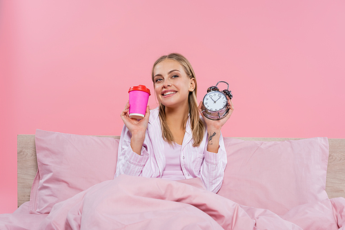 Positive blonde woman in pajama holding alarm clock and coffee to go isolated on pink