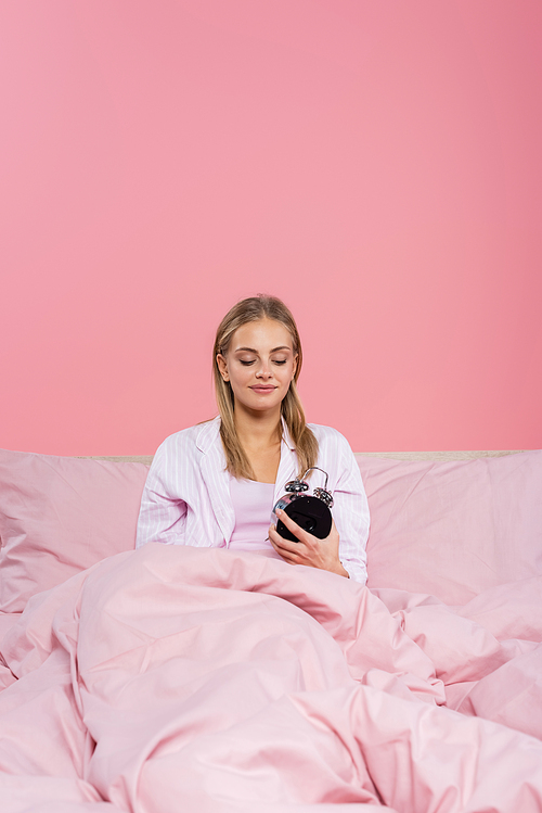 Young woman looking at alarm clock on bed isolated on pink