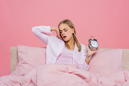 Blonde woman in pajamas holding alarm clock and yawning on bed isolated on pink