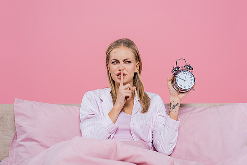 Blonde woman showing secret gesture and holding alarm clock on bed isolated on pink