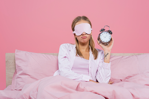 Sad woman in sleep mask holding alarm clock on bed isolated on pink