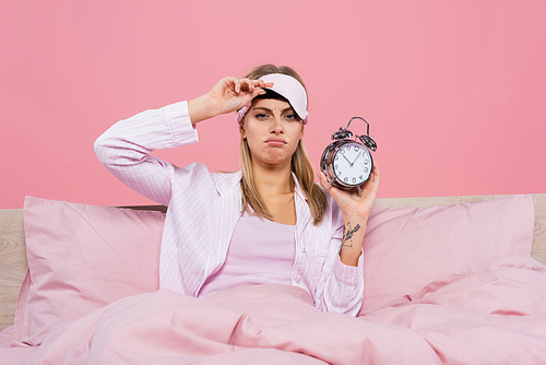 Upset woman holding sleep mask and alarm clock on bed isolated on pink