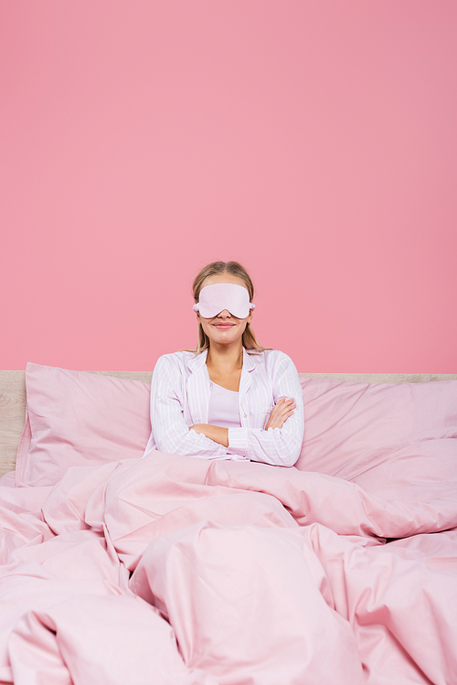 Smiling woman in sleep mask sitting with crossed arms on bed isolated on pink