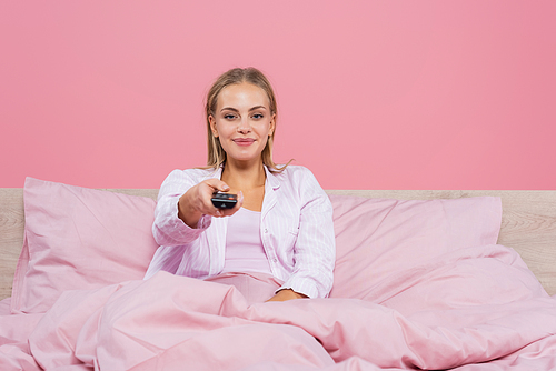 Smiling woman in pajama holding remote controller on bed isolated on pink