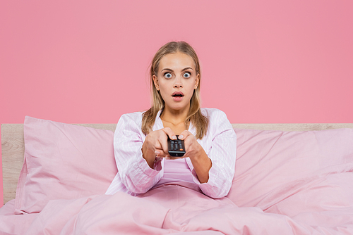 Shocked blonde woman in pajamas holding remote controller on bed isolated on pink