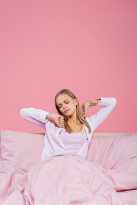 Young blonde woman in pajamas stretching on bed isolated on pink