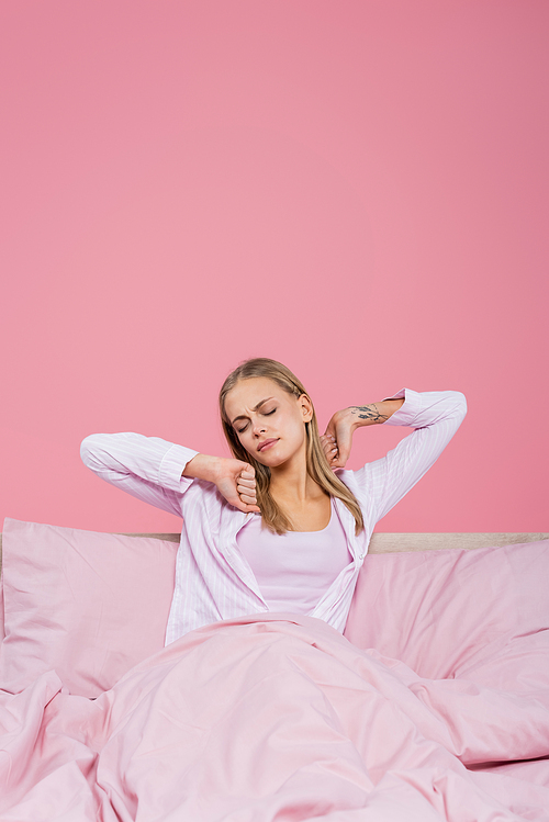 Young blonde woman in pajamas stretching on bed isolated on pink