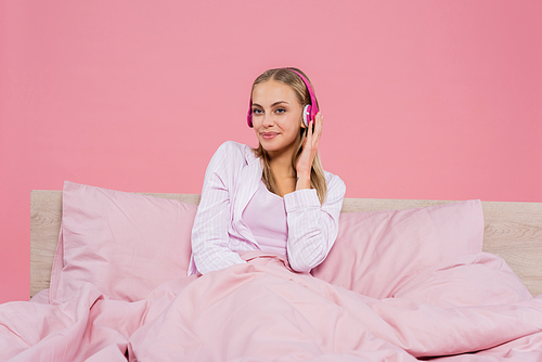 Young blonde woman in pajamas listening music in headphones on bed isolated on pink