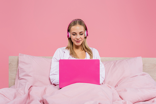 Smiling freelancer in headphones using laptop on bed isolated on pink