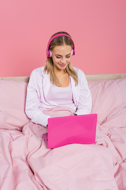 Positive woman in headphones and pajamas using laptop on bed isolated on pink