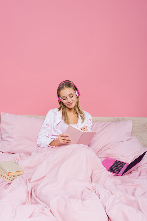 Freelancer in headphones writing on notebook near laptop and books on bed isolated on pink