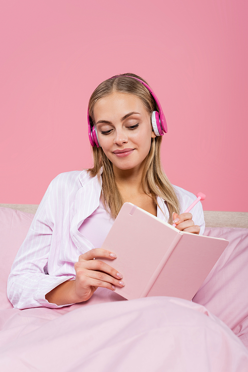 Smiling blonde woman in headphones writing on notebook on bed isolated on pink