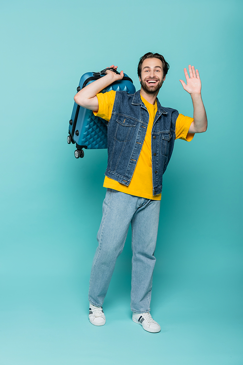 Happy traveler in denim vest holding suitcase and waving hand on blue background