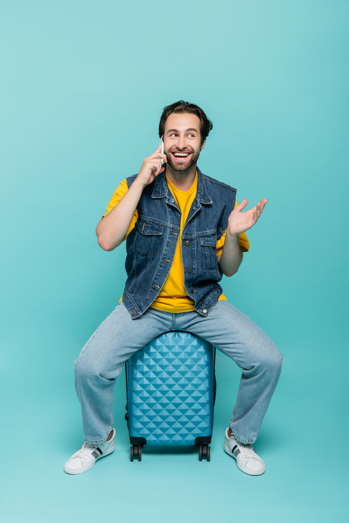 Smiling man talking on cellphone while sitting on suitcase on blue background