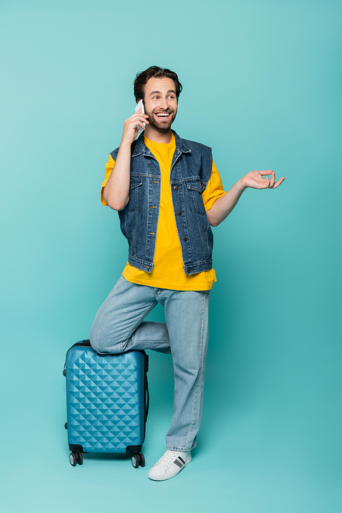 Happy tourist in denim vest talking on smartphone near baggage on blue background