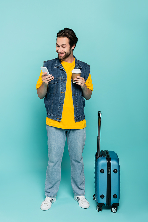 Smiling man with coffee to go using smartphone near suitcase on blue background