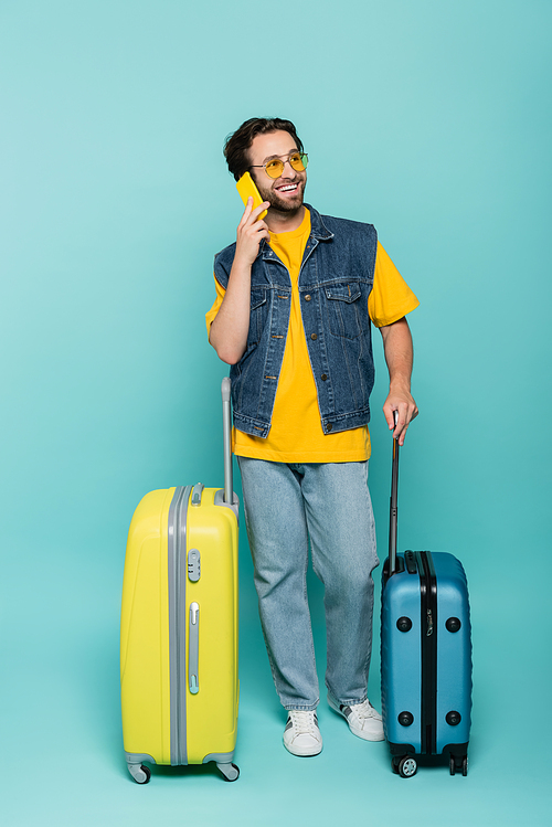 Smiling man in sunglasses talking on smartphone near suitcases on blue background