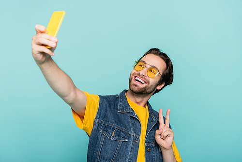 Smiling man in denim vest showing peace sign while taking selfie isolated on blue