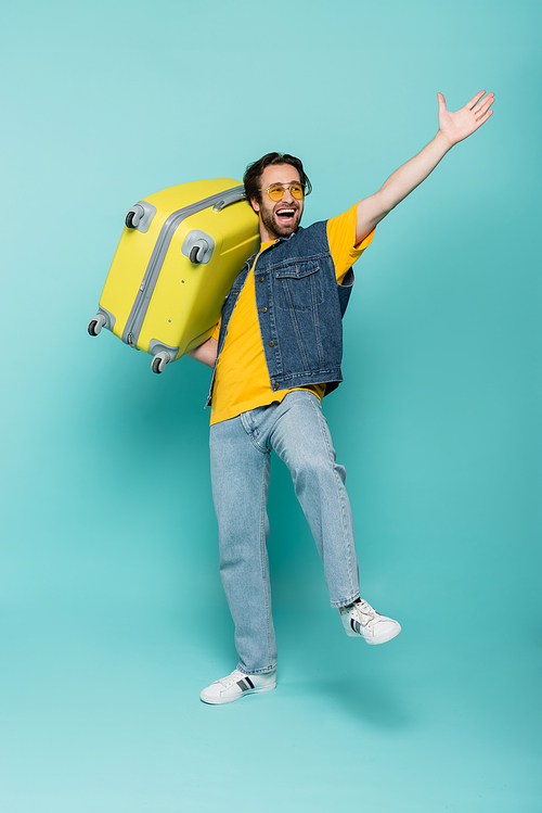 Excited tourist in sunglasses holding suitcase on blue background
