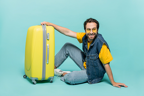 Smiling man in sunglasses sitting near suitcase on blue background