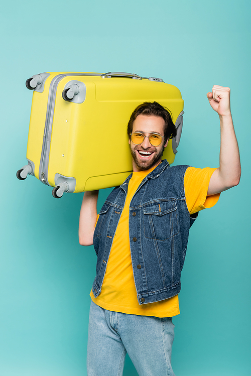 Cheerful man in sunglasses showing yes gesture while holding suitcase isolated on blue