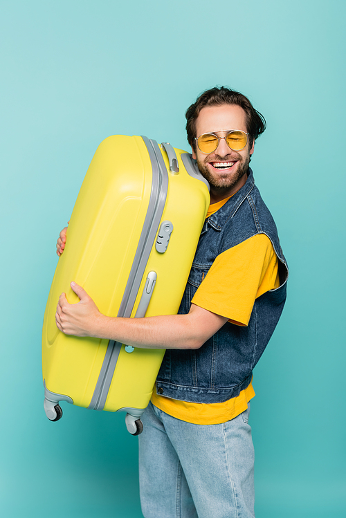 Happy man in sunglasses hugging suitcase isolated on blue