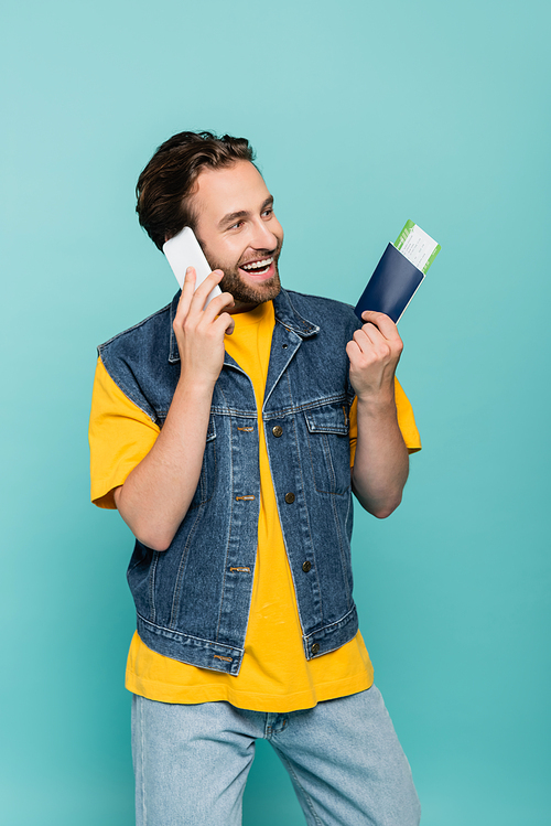 Cheerful tourist with passport talking on smartphone isolated on blue